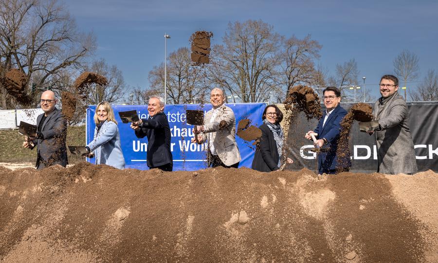 Spatenstich für das neue Parkhaus Unterer Wöhrd: Sieben Personen schaufeln Sand vom vorbereiteten Sandhügel auf dem Baugelände.