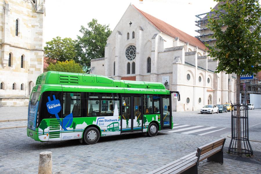 Grüner Elektrobus fährt an Dom und Ulrichskirche vorbei, umgeben von Bäumen und Kopfsteinpflasterstraße.
