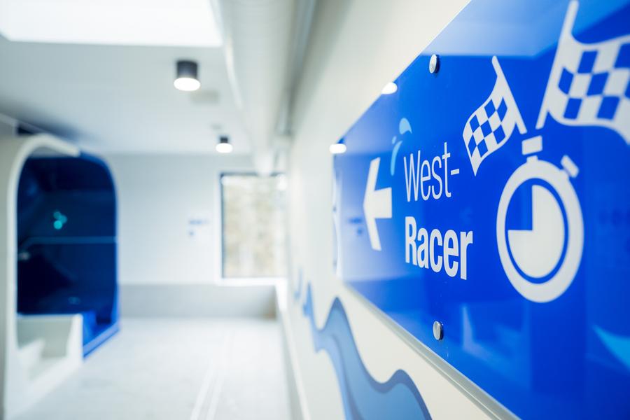 Blaues Wegweiser-Schild „West-Racer“ mit Pfeil in einem hellen Innenbereich.