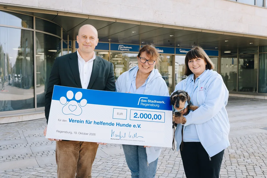 Von links: Mathis Broelmann, kaufmännischer Leiter von das Stadtwerk.Regensburg, übergibt einen Spendenscheck über 2.000 Euro an Gabi Feneberg und Birgit Forster vom Verein für helfende Hunde e.V. Birgit Forster hält den Vorlesehund „Franzl“ auf dem Arm. Die Übergabe findet vor einem Gebäude von das Stadtwerk.Regensburg statt.