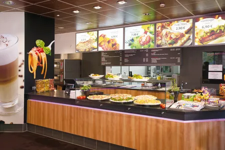 Theke mit verschiedenen Pizzen und Salaten in einem Restaurant, darüber digitale Menüs mit Preisen für Pizza, Hamburger und Snacks