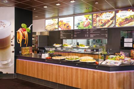 Theke mit verschiedenen Pizzen und Salaten in einem Restaurant, darüber digitale Menüs mit Preisen für Pizza, Hamburger und Snacks