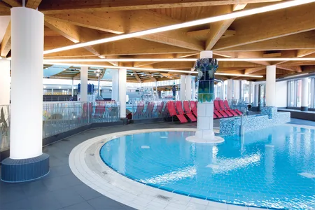 Innenbereich eines Schwimmbads mit rundem Becken, Holzdecke und roten Liegestühlen am Beckenrand