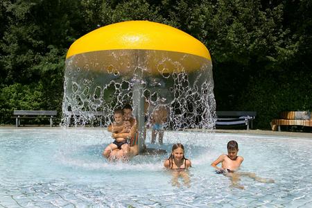 Kinder spielen im flachen Wasser eines Planschbeckens unter einem großen gelben Pilz-Wasserspiel mit herabfallendem Wasser