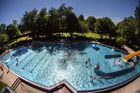 Außen-Schwimmbecken mit mehreren Schwimmern, Wasserrutsche rechts, umgeben von Bäumen und Wiese bei sonnigem Himmel