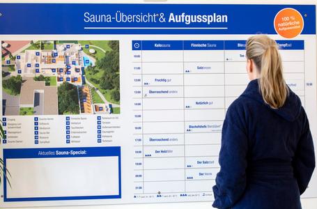 Eine blonde Person im Bademantel steht vor dem Sauna-Aufgussplan.