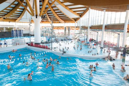 Innenansicht des Westbads mit mehreren Schwimmbecken, vielen Schwimmern und großen Fenstern unter einer Holzdecke