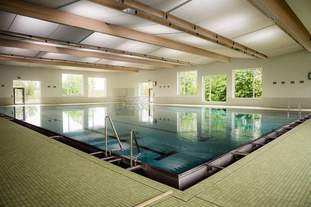 Innenansicht eines Schwimmbads mit großem Becken, Treppenstufen und großen Fenstern mit Blick auf Bäume
