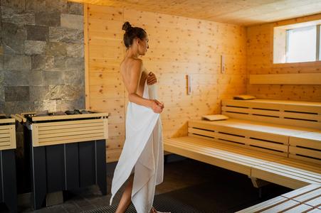 Frau mit hochgestecktem Haar, die ein weißes Handtuch hält, steht in einer Sauna mit Holzwänden und Holzbänken neben einem Saunaofen