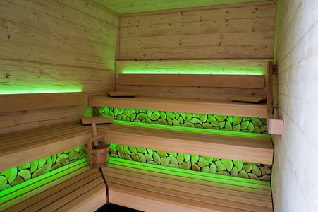 Innenraum einer Sauna mit Holzbänken und grün beleuchteten Holzscheiten unter den Sitzflächen