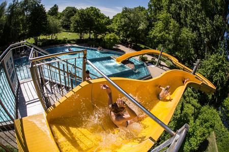 Kinder rutschen eine gelbe Wasserrutsche im Freibad hinunter, umgeben von Bäumen und Wiesen