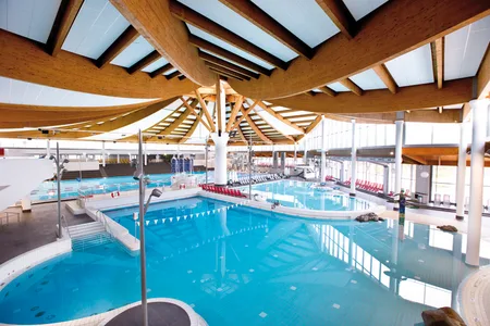 Innenansicht eines großen Hallenbads mit mehreren Schwimmbecken und sichtbaren Holzträgern an der Decke