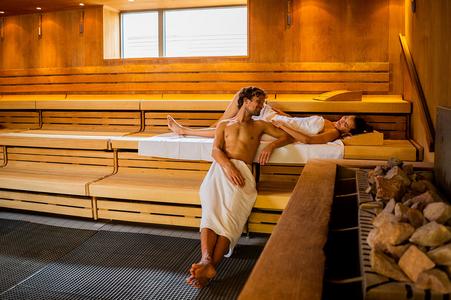 Zwei Personen entspannen in einer Holzsauna, eine liegt auf der oberen Bank, die andere sitzt auf der unteren Bank mit einem Handtuch um die Hüften