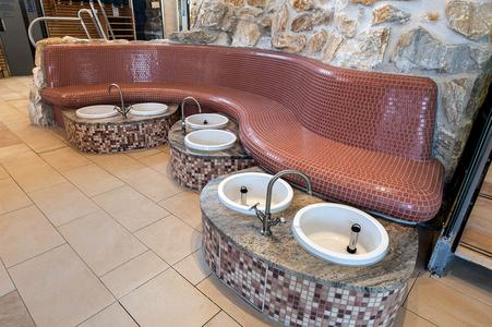 Geschwungene Sitzbank mit roten Mosaikfliesen und drei Fußwaschbecken mit Wasserhähnen vor einer Natursteinwand