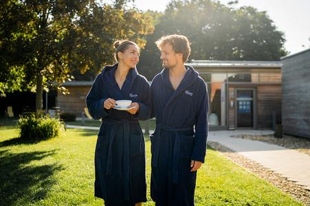 Zwei Personen in blauen Bademänteln stehen auf einer Wiese vor einem modernen Gebäude, eine hält eine Tasse mit Untertasse
