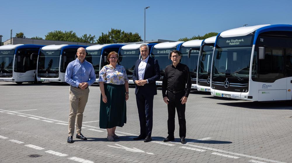 Vier Personen stehen auf dem Betriebshof vor einer Reihe moderner weiß-blauer Elektrobusse von das Stadtwerk.Mobilität. Die Fahrzeuge sind nebeneinander aufgestellt.