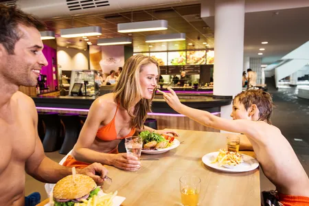 Drei Personen in Badebekleidung sitzen an einem Tisch in einem Restaurant, ein Kind reicht einer Frau Essen, auf dem Tisch Teller mit Burger, Pommes und Salat