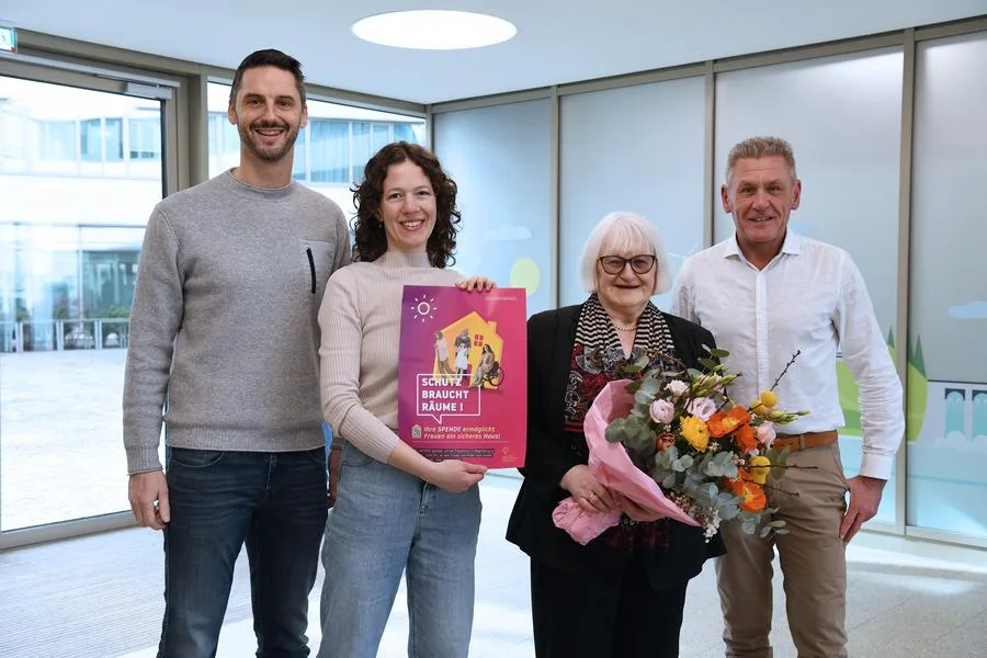 Vier Personen stehen nebeneinander im Foyer des Verwaltungsgebäudes, einem Raum mit großen Fenstern, und lächeln in die Kamera. Karin Roth hält ein pinkes Plakat mit der Aufschrift „Schutz braucht Räume!“, daneben steht Margit Kunc, die einen bunten Blumenstrauß hält. Dies ist die offizielle Spendenübergabe für das soziales Projekt "Frauen helfen Frauen e.V."