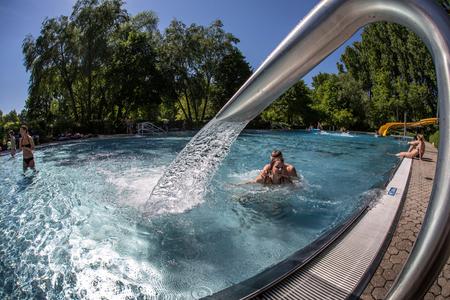 Wasserstrahl aus Metallrohr in ein Schwimmbecken mit mehreren Personen und Bäumen im Hintergrund