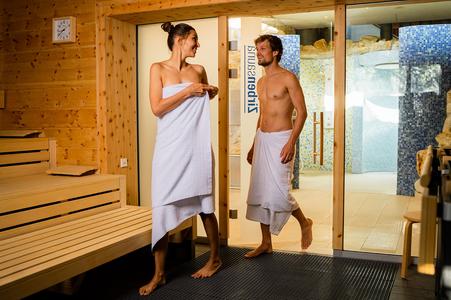 Zwei Personen in einem Saunabereich, beide mit weißen Handtüchern bekleidet, stehen vor einer Glastür mit der Aufschrift 'Zirbensauna'