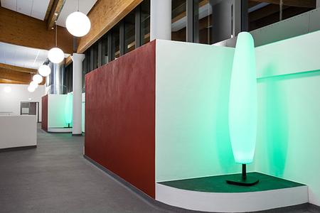 Innenraum mit moderner Beleuchtung, weißen Säulen und einer grünen, leuchtenden Stehlampe neben einer roten Wand