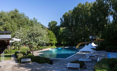 Leeres Freibad mit klarem Wasser, Wasserrutsche und Liegestühlen, umgeben von Bäumen unter blauem Himmel