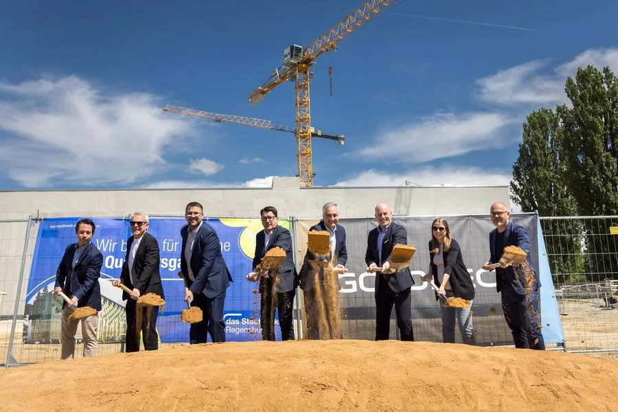 Mehrere Personen stehen nebeneinander auf einer Baustelle und werfen beim symbolischen Spatenstich Sand nach vorne; im Hintergrund sind ein Baukran, Bauzäune und ein Banner mit Bezug zu einem Bauprojekt in Regensburg zu sehen.