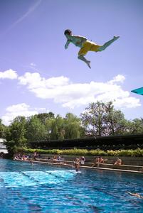 Mann in gelber Badehose springt vom Sprungturm in ein 50-Meter-Schwimmbecken bei sonnigem Wetter