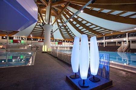 Innenansicht des Westbads mit beleuchteten Säulen, Holztragwerk und Schwimmbecken im Hintergrund