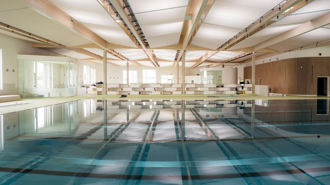 Innenansicht eines großen Hallenbads mit Schwimmbecken und Holzdeckenbalken