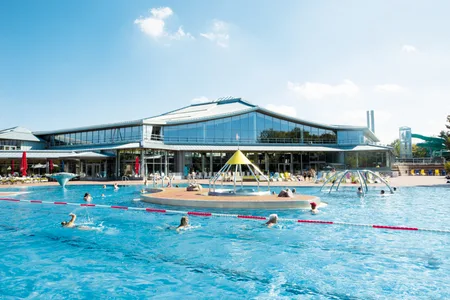 Außenbecken des Westbads mit Schwimmern und Wasserspielen vor moderner Halle unter blauem Himmel