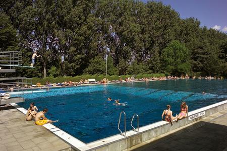 50-Meter-Schwimmbecken mit Sprungturm links, Menschen schwimmen und sonnen sich, Bäume im Hintergrund