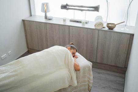 Person liegt auf Massageliege, bedeckt mit weißer Decke, in hellem Raum mit Holzschrank und Wellnessutensilien