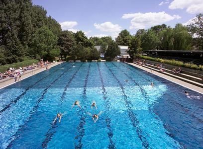 50-Meter-Freiluft-Schwimmbecken mit mehreren Schwimmern und Liegewiesen an beiden Seiten unter blauem Himmel