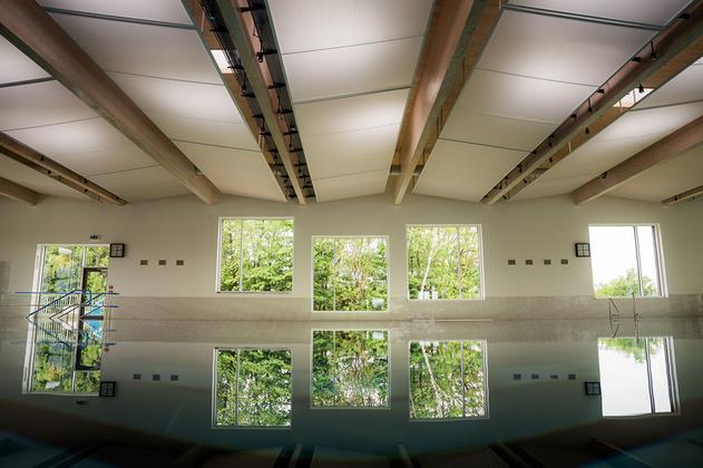 Innenansicht eines Schwimmbeckens mit großen Fenstern, die Bäume draußen zeigen, und sichtbaren Deckenbalken