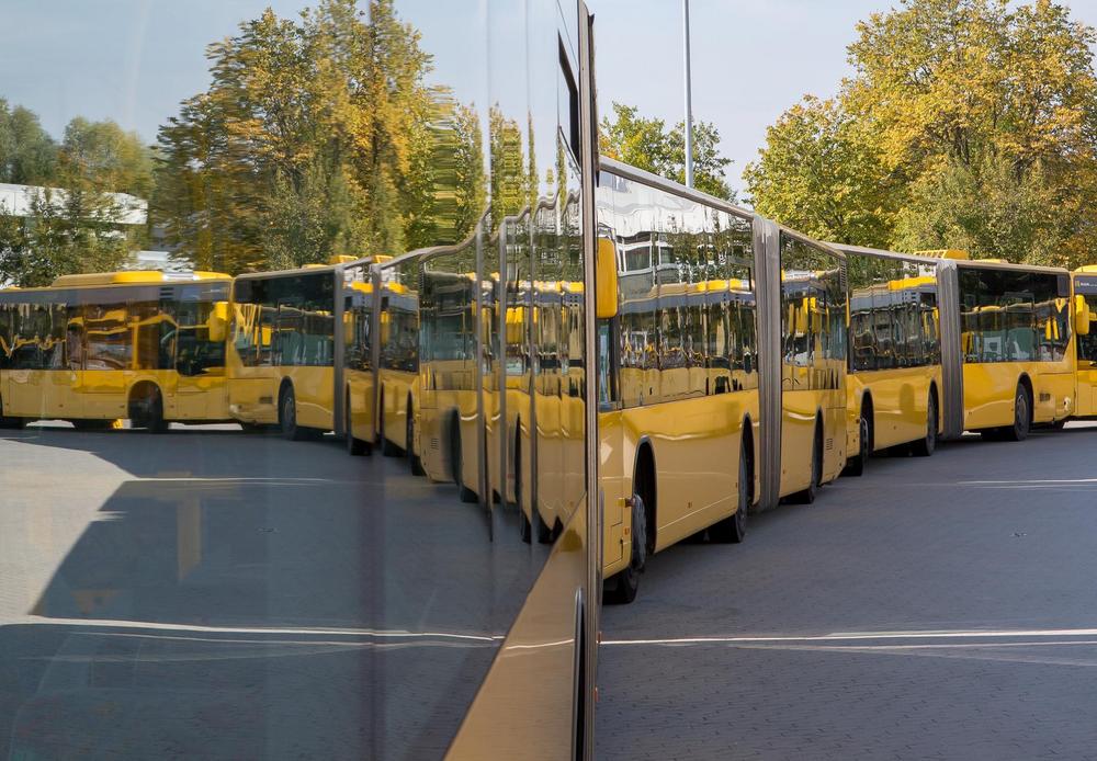 Mehrere gelbe Gelenkbusse stehen auf einem Betriebshof. Die spiegelnde Seitenfläche eines Busses reflektiert die umliegenden Fahrzeuge und erzeugt einen symmetrischen, nahezu kaleidoskopartigen Effekt. 