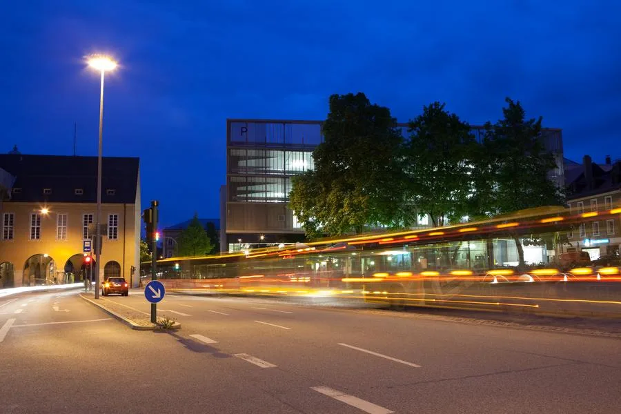Aufnahme einer Stadtstraße bei Dunkelheit. Ein vorbeifahrender Bus erzeugt durch die Langzeitbelichtung leuchtende Lichtspuren.