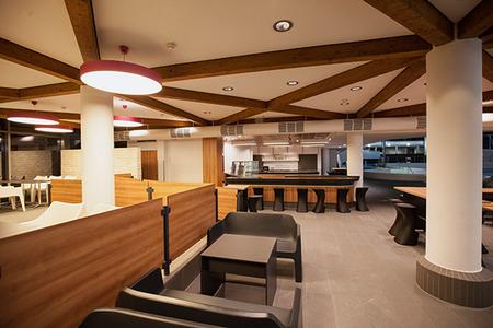 Innenraum eines modernen Cafés mit Holzdeckenbalken, Sitzbereichen aus schwarzen Sesseln und Holztischen, Theke im Hintergrund