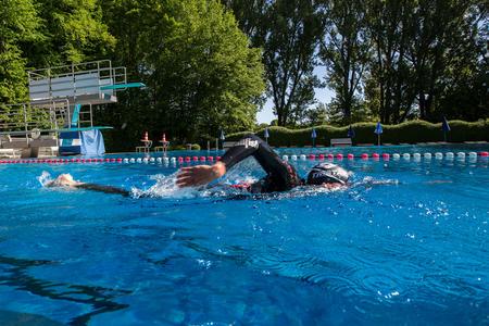 Schwimmer in schwarzem Neoprenanzug und Schwimmbrille beim Kraulschwimmen im Freibad mit Sprungturm und Bäumen im Hintergrund