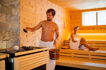 Mann in Sauna gießt Wasser mit Kelle auf heiße Steine, Frau sitzt entspannt auf Holzbänken