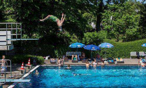 Junger Mann macht einen Rückwärtssalto vom Sprungbrett in ein Schwimmbecken mit mehreren Schwimmern und Sonnenschirmen am Beckenrand