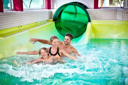 Drei Personen im Schwimmbadbecken am Ende einer grünen Wasserrutsche, umgeben von weißen Fliesen
