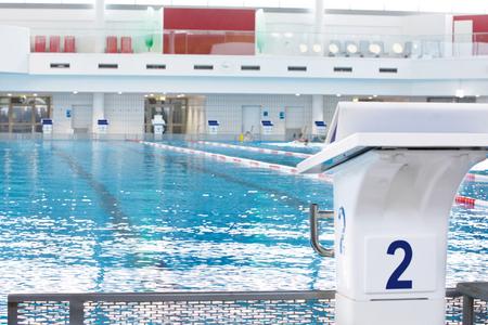 Startblock Nummer 2 am Beckenrand eines Hallenbads mit Schwimmerbahnen und Liegestühlen im Hintergrund