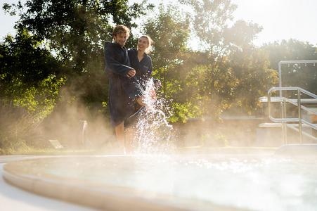 Zwei Personen in Bademänteln stehen am Rand eines dampfenden Außenpools, eine Person spritzt Wasser mit dem Fuß