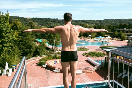 Mann in schwarzer Badehose steht mit ausgestreckten Armen auf einem Sprungbrett über einem Schwimmbecken im Freien