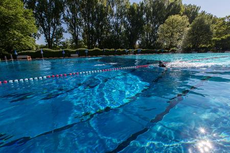 Schwimmer im Außenbecken eines Freibads, umgeben von Bäumen und Sonnenschirmen am Beckenrand