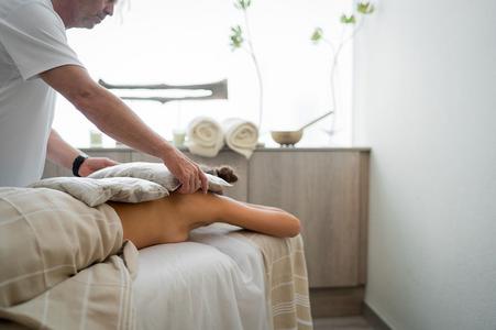 Mann legt Kissen auf den Rücken einer Person, die in eine Decke gewickelt auf Massageliege in einem hellen Raum liegt