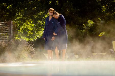 Zwei Personen in blauen Bademänteln stehen an einem dampfenden Außenpool, umgeben von Bäumen und Sonnenlicht