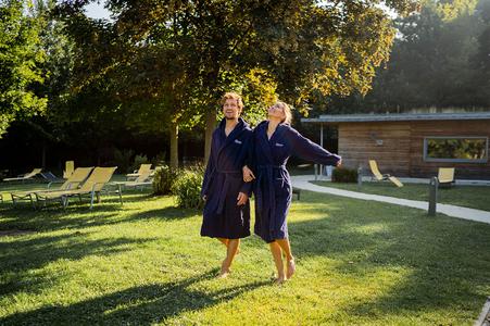 Zwei Personen in dunkelblauen Bademänteln gehen barfuß auf einer Wiese neben einem Holzgebäude mit Liegestühlen im Hintergrund