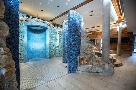 Große Erlebnisdusche mit blauen Mosaikfliesen und Natursteinwänden in einem Wellnessbereich mit Holzverkleidung und hellem Boden